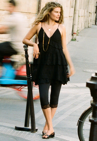 A woman stands on a city sidewalk wearing a black sleeveless ruffled mini dress with thin straps, black capri leggings with lace trim, and black flat sandals. She is accessorized with layered gold necklaces.