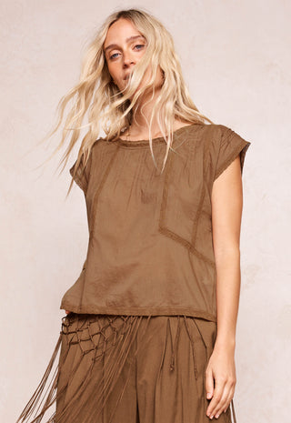 a-woman-with-long-blonde-hair-wearing-a-brown-short-sleeve-top-and-matching-brown-fringe-skirt-posing-against-a-neutral-beige-background