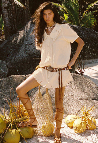 a-woman-stands-outdoors-among-rocks-palm-leaves-and-coconuts-wearing-a-loose-white-short-sleeve-blouse-and-matching-white-skirt-or-shorts-styled-with-a-brown-belt-brown-lace-up-sandals-and-a-woven-bag